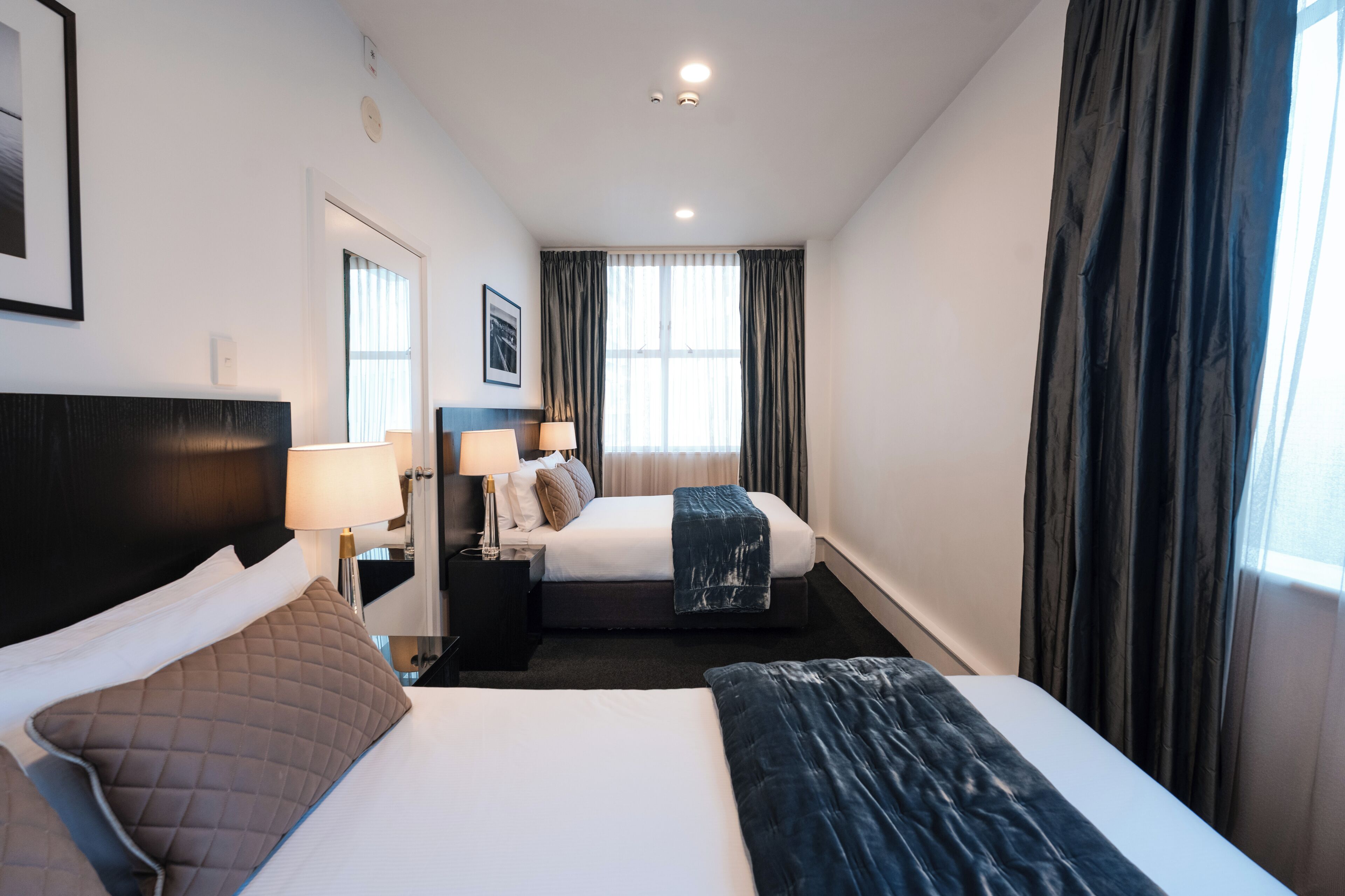A hotel room with a large bed, a smaller bed, a nightstand with a lamp, a framed picture on the wall, and a window with curtains.