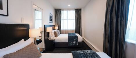 Habitación de hotel con una cama grande, otra más pequeña, una mesita de noche con lámpara, un cuadro en la pared y una ventana con cortinas.