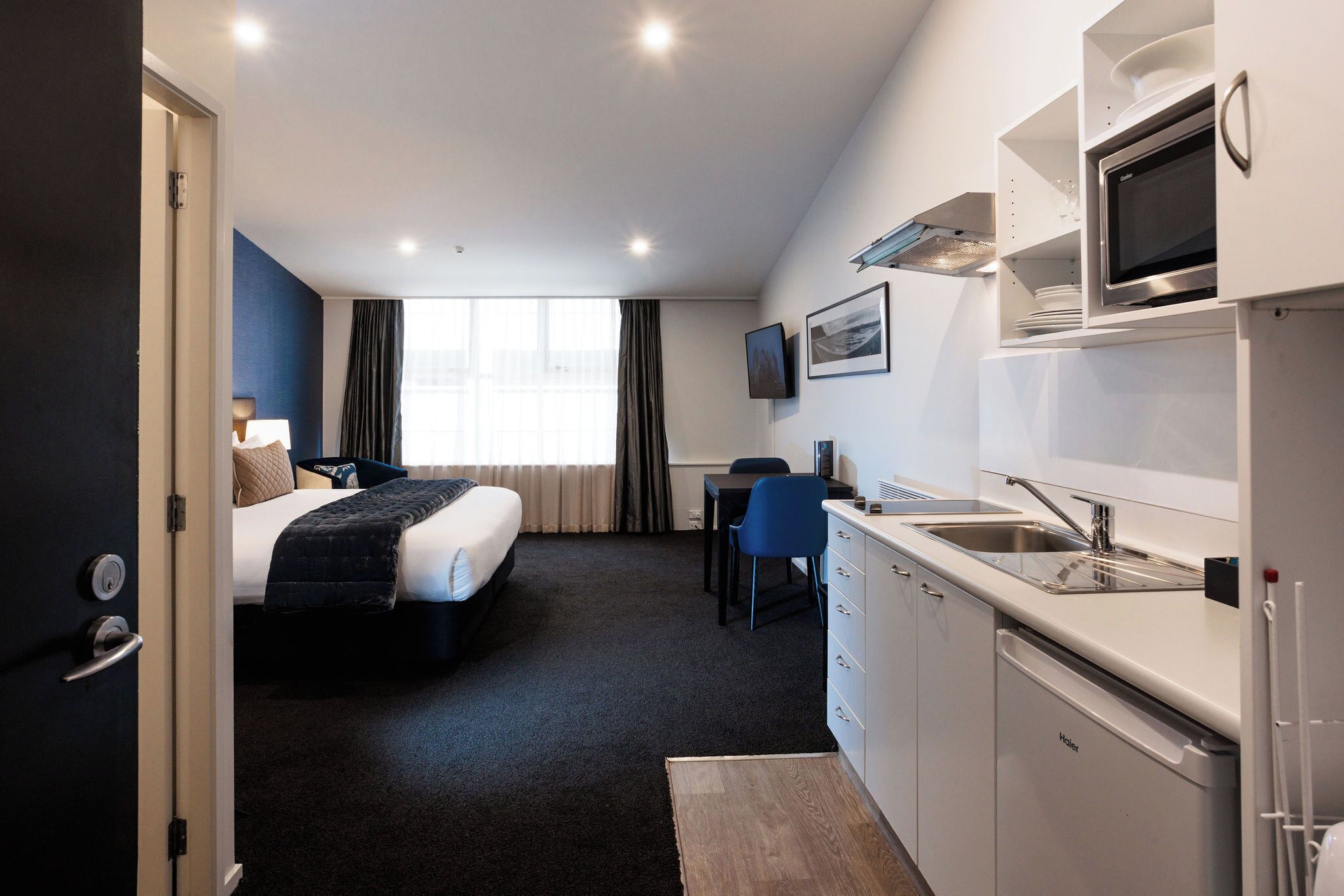 A hotel room with a bed, a desk, a chair, a kitchenette, and a TV.