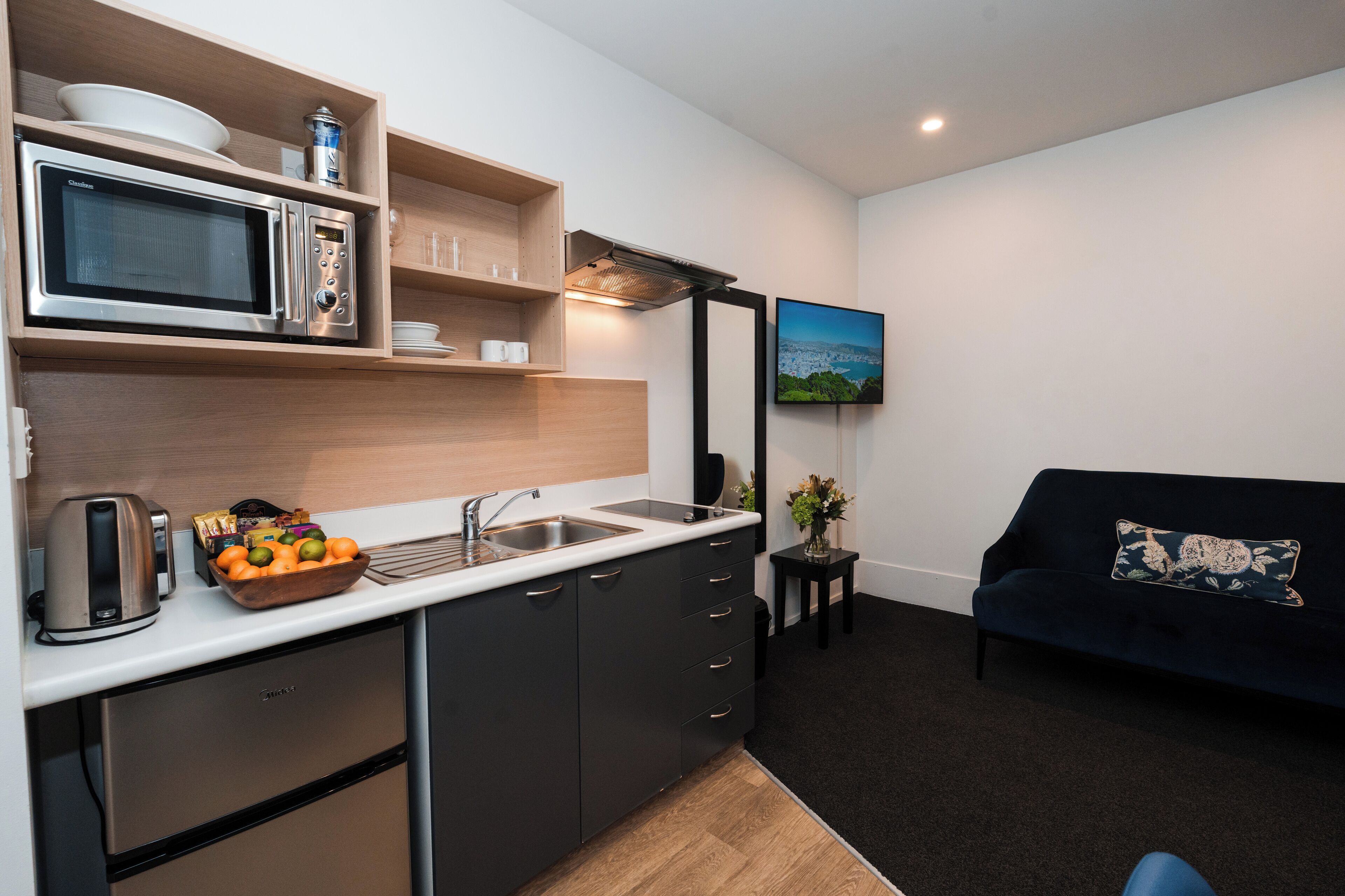 A compact kitchen with a microwave, kettle, and fruit bowl. There's a sofa and a small table with a plant.