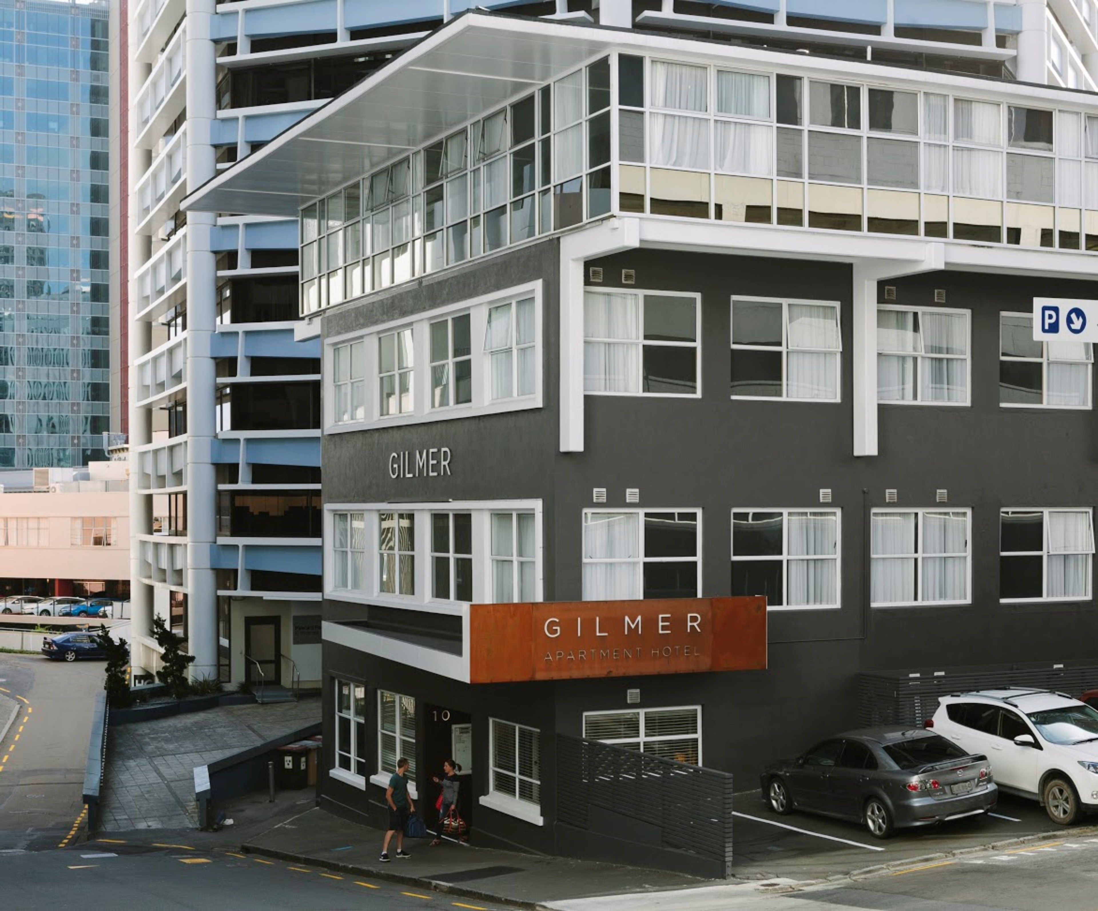 A modern apartment hotel named 'GILMER' with a prominent sign, multiple floors, and a parking area.