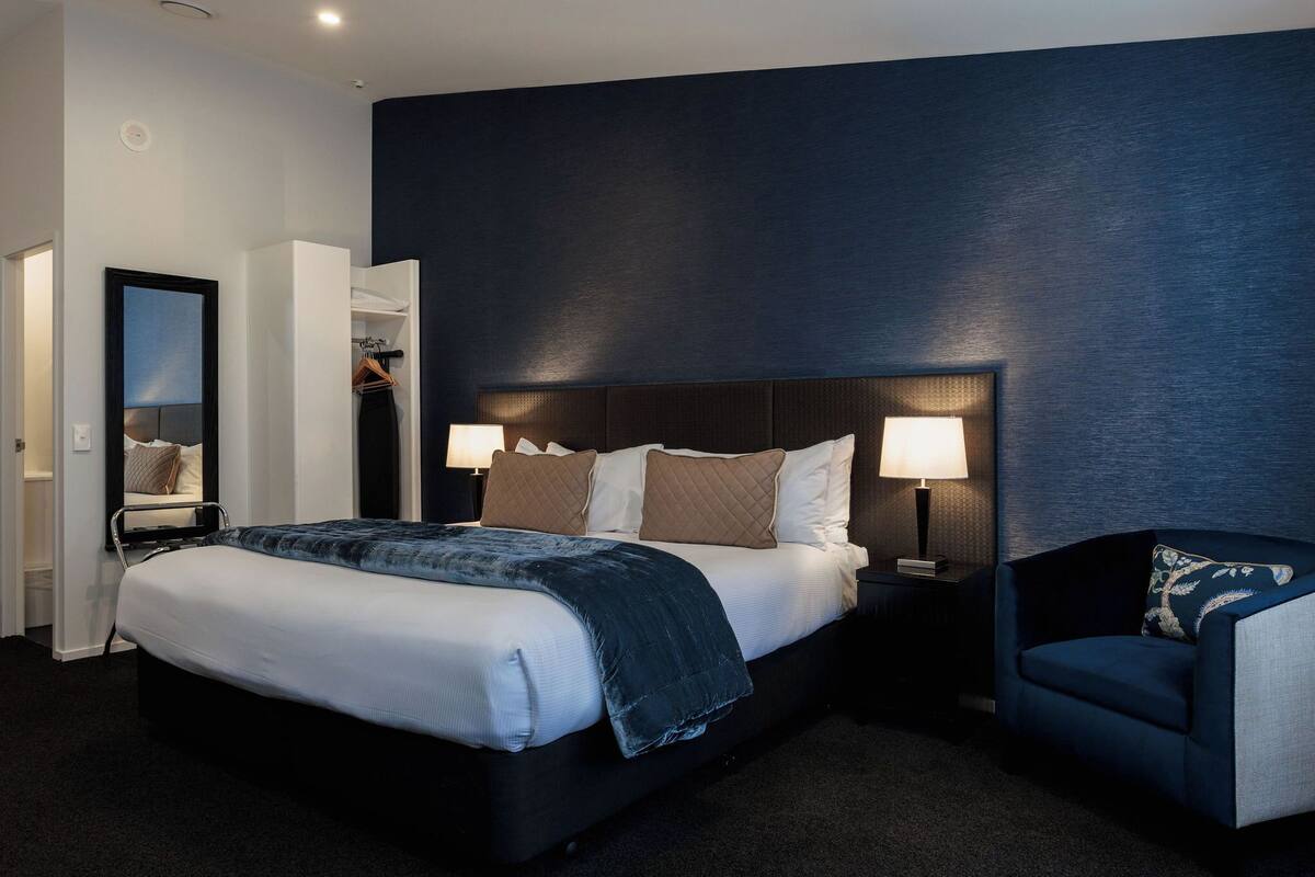 Una habitación de hotel moderna con una cama grande, lámparas de noche, una pared de acento azul oscuro y una zona de estar con una silla.