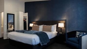 A modern hotel room with a large bed, bedside lamps, a dark blue accent wall, and a seating area with a chair.