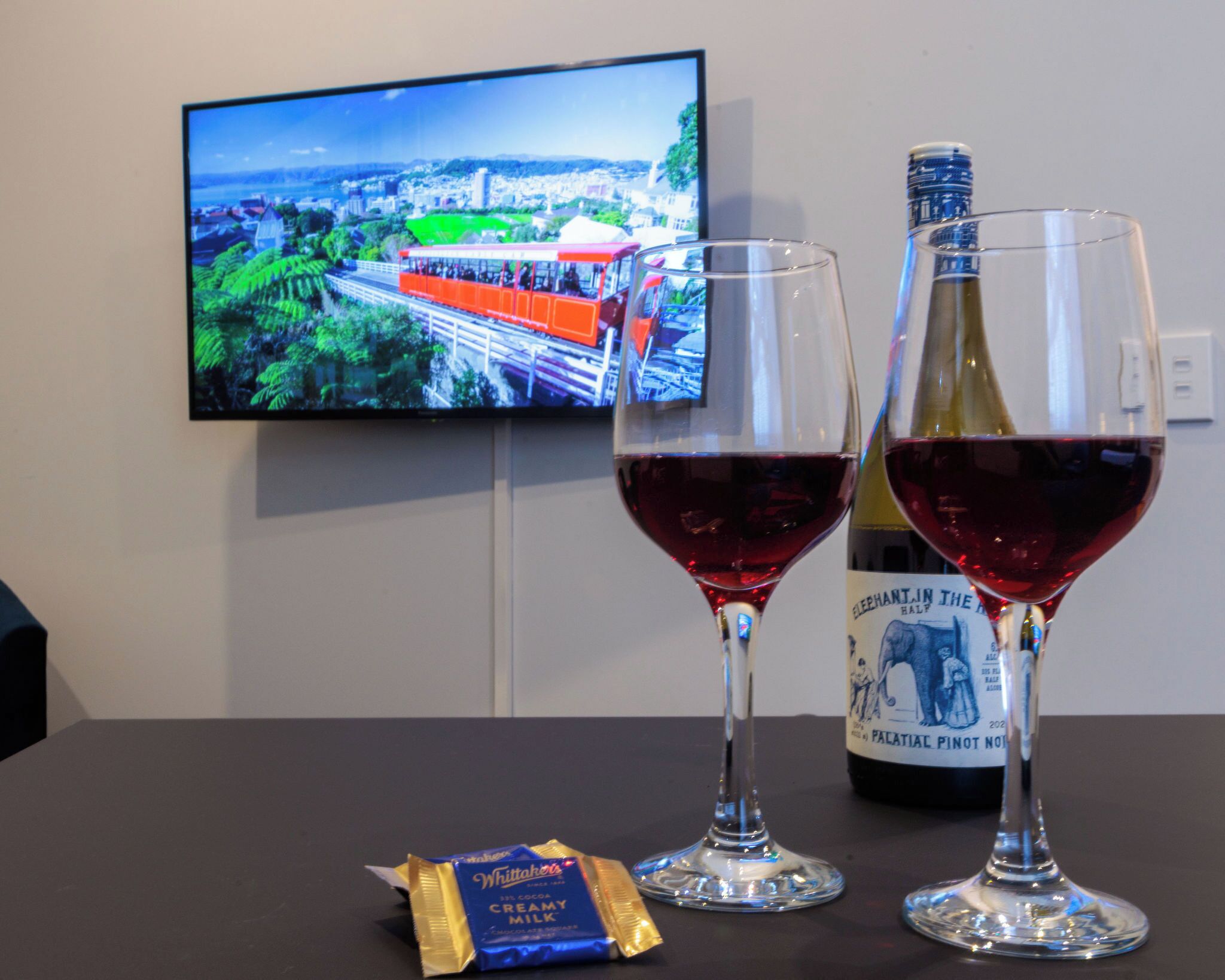 Two wine glasses on a table with a bottle of wine and a chocolate bar. A television displaying a scenic view is mounted on the wall behind.