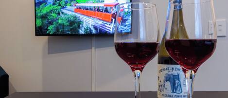 Two wine glasses on a table with a bottle of wine and a chocolate bar. A television displaying a scenic view is mounted on the wall behind.