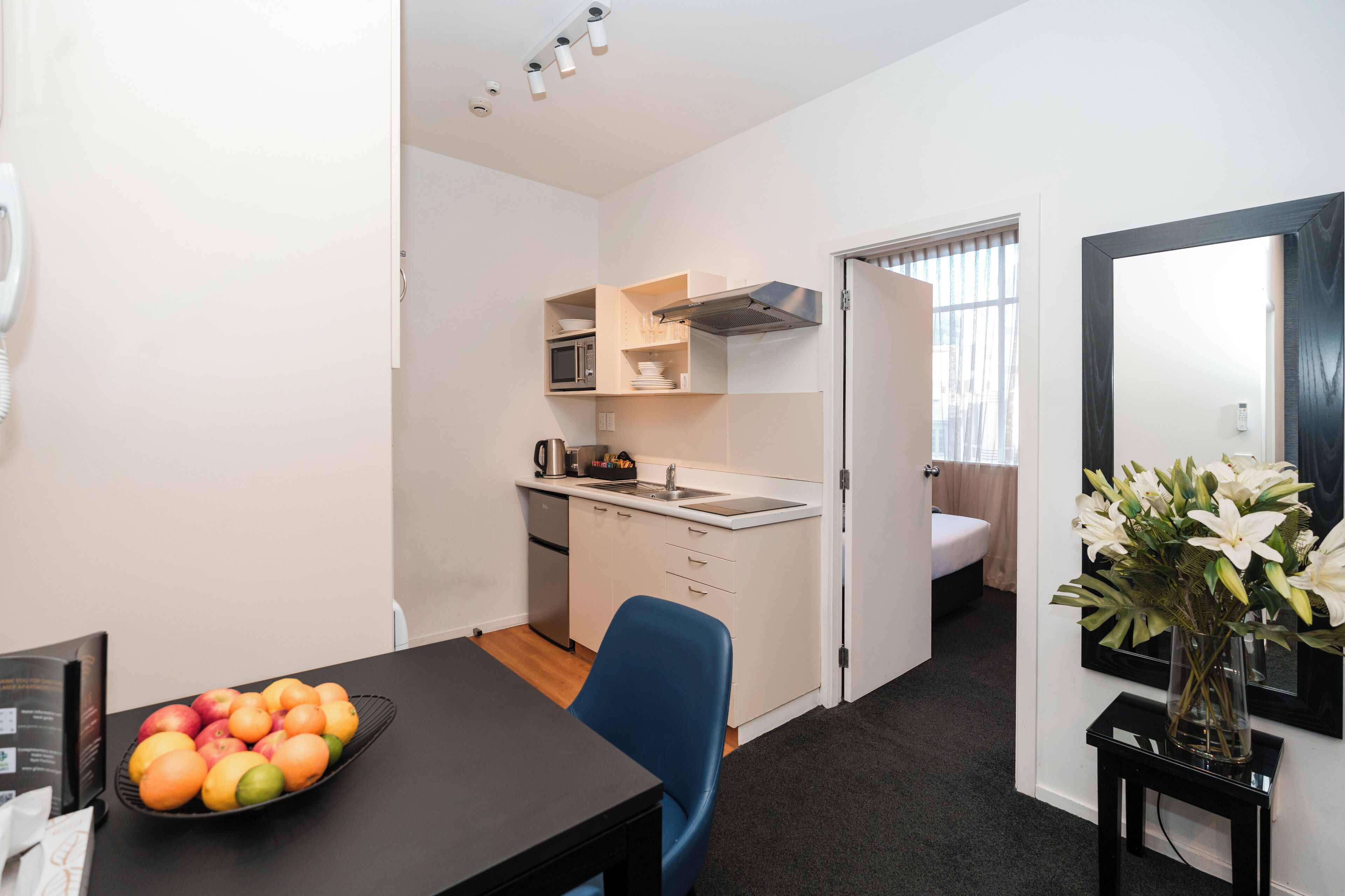 Habitación de hotel compacta con una mesa de comedor, una silla azul, un frutero, una kitchenette, un baño y un espejo con un florero.
