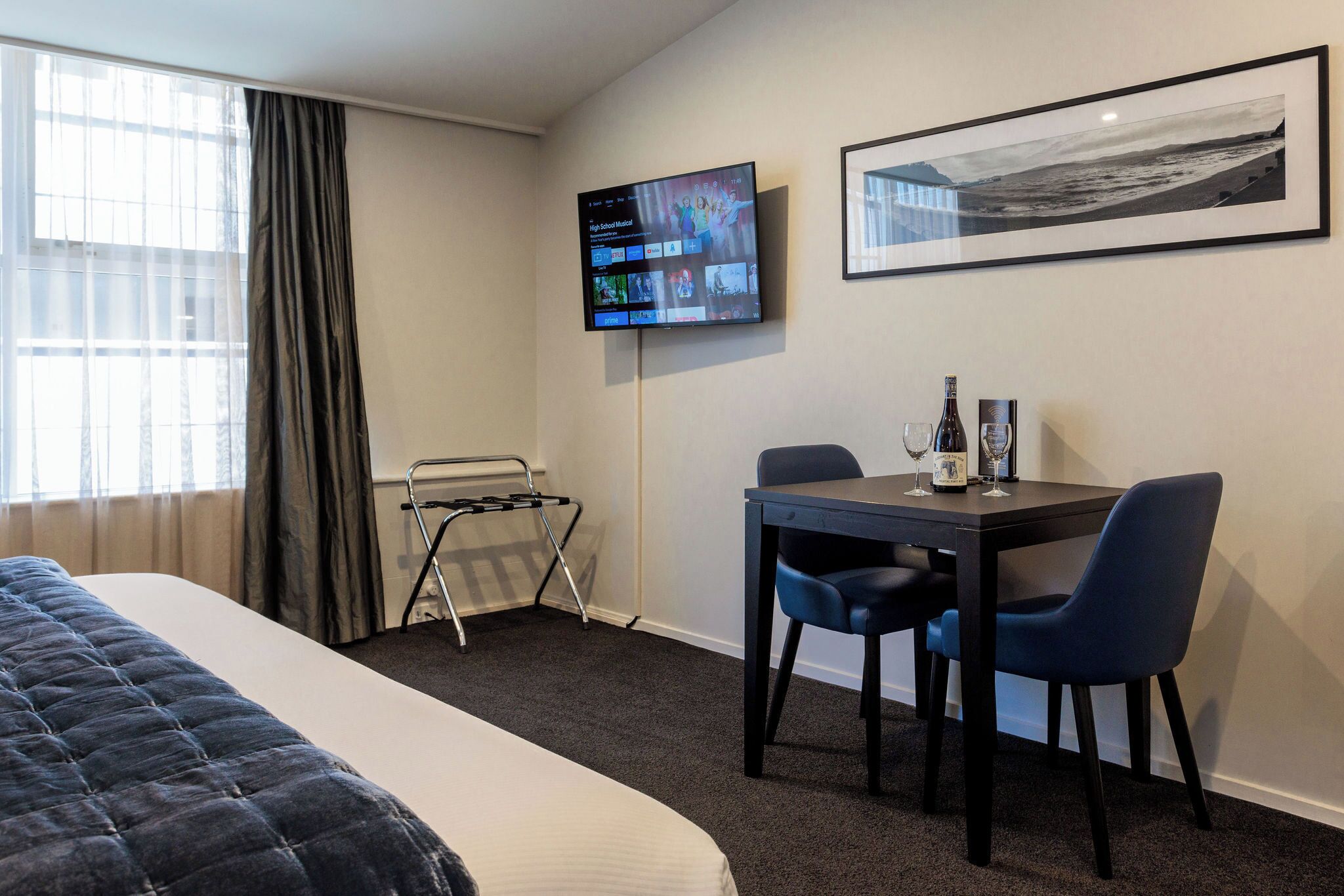 A hotel room with a bed, a desk with two chairs, a television mounted on the wall, and a framed picture.