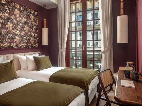 A hotel room with two beds, a large window with curtains, a wooden chair, a table with mugs, and a wall with a floral pattern.