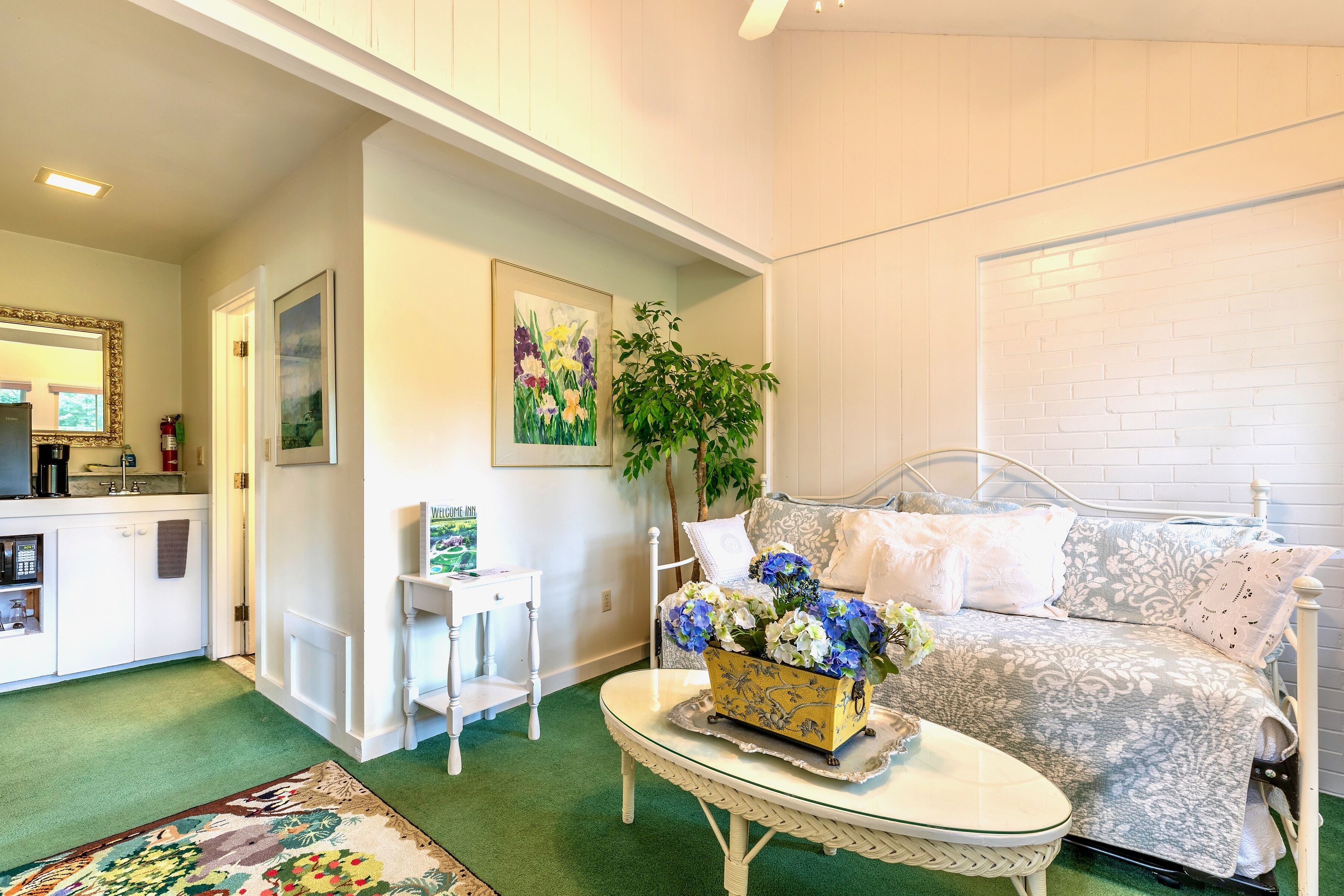 Una sala de estar acogedora con alfombra verde, un sofá blanco con cojines estampados, una mesita blanca con un jarrón de flores y un cuadro en la pared.