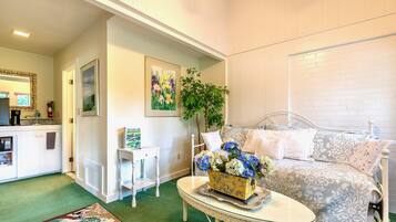 Una sala de estar acogedora con alfombra verde, un sofá blanco con cojines estampados, una mesita blanca con un jarrón de flores y un cuadro en la pared.