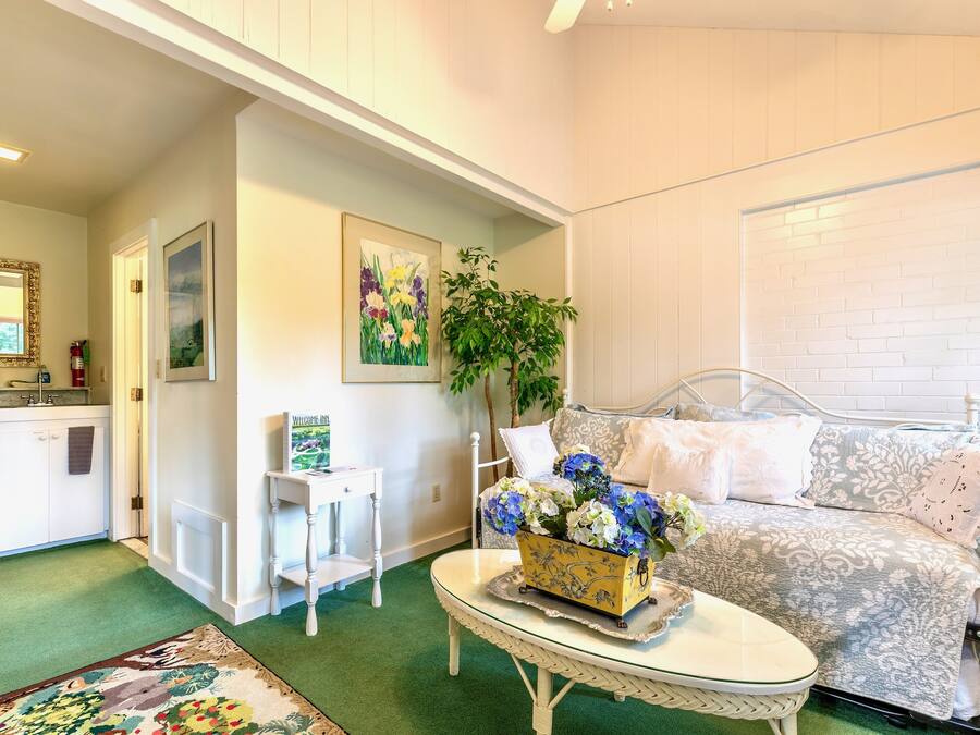 Una sala de estar acogedora con alfombra verde, un sofá blanco con cojines estampados, una mesita blanca con un jarrón de flores y un cuadro en la pared.