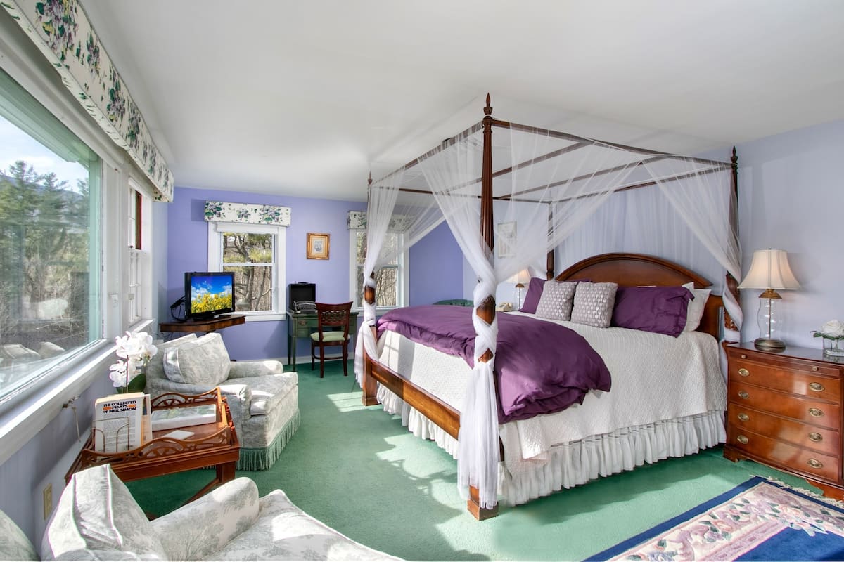 Ein Bett mit Baldachin, bezogen mit einem violetten Überwurf, ein Nachttisch aus Holz, ein Schreibtisch mit Computer und ein Fenster mit Blick auf Bäume.