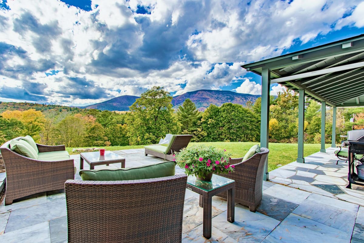 Eine Terrasse im Freien mit Korbmöbeln, einem Glastisch und Blick auf Berge und Bäume.