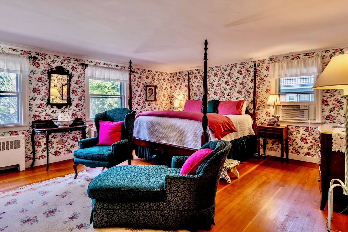 Ein Schlafzimmer mit floralem Wandpapier, einem Himmelbett, einem Nachttisch, einem Sessel und einem Fenster mit Vorhängen.