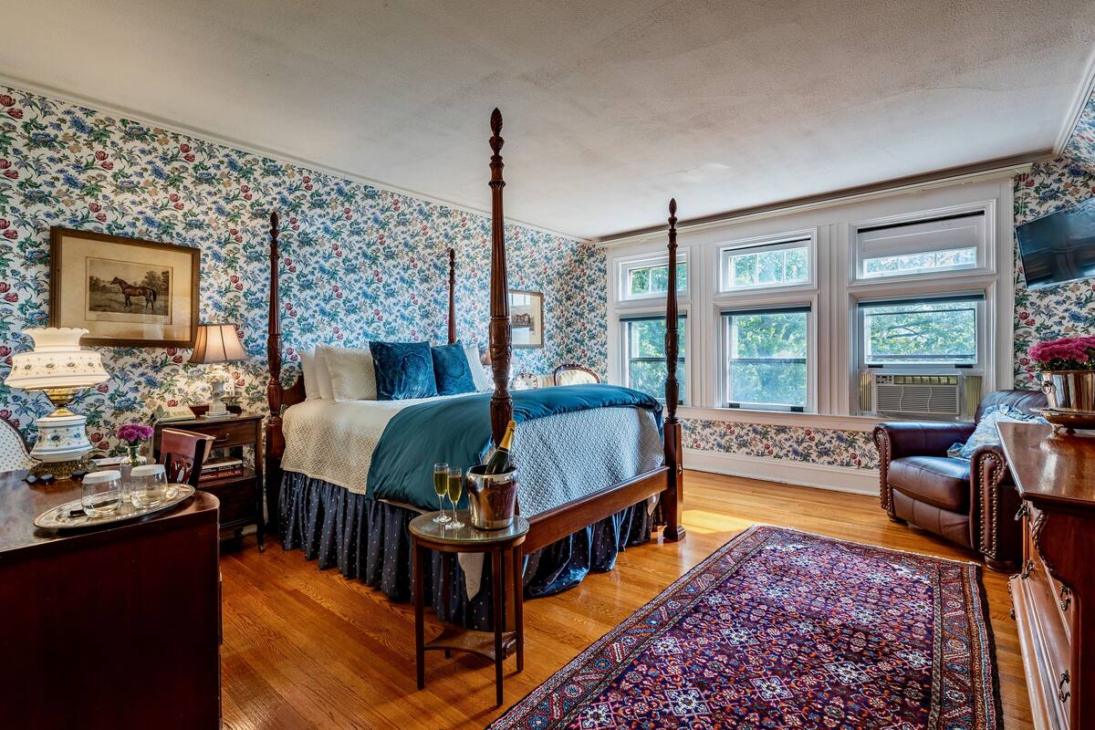 Ein Schlafzimmer mit einem Himmelbett, floralem Wandpapier, einem großen Fenster und Holzboden.