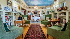 A cozy room with green upholstered chairs, a wooden coffee table, and bookshelves filled with books. A red patterned rug is on the floor, and a chandelier hangs from the ceiling.