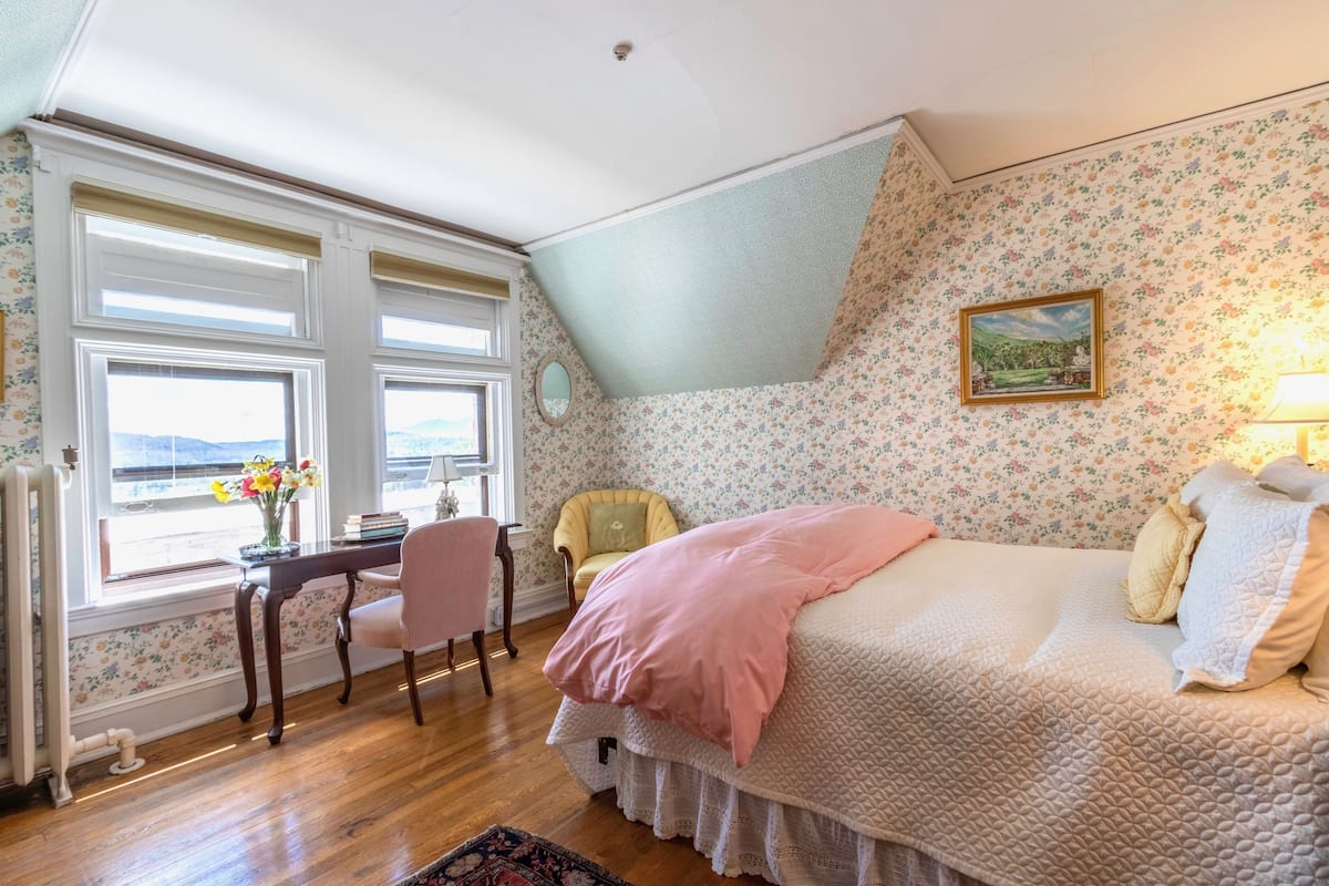 Ein Schlafzimmer mit Bett, Schreibtisch, Stuhl, Heizkörper und einem Fenster mit Blick.