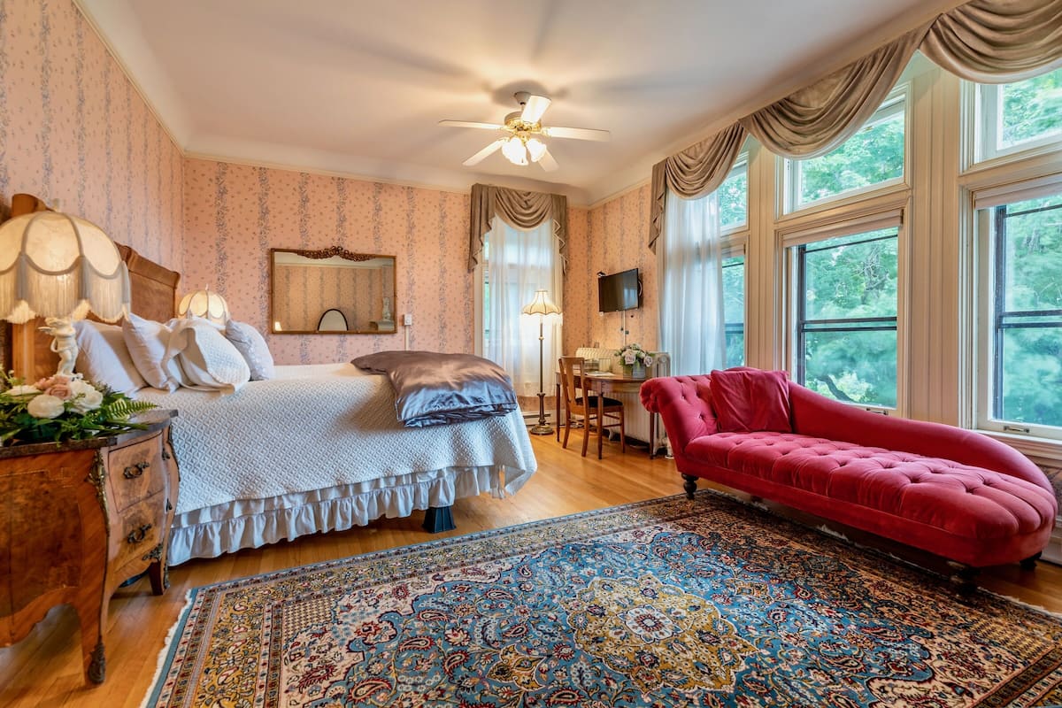 Ein Schlafzimmer mit einem Bett, einem roten Sofa, einem Schreibtisch, einem Sessel, einem Fernseher und großen Fenstern.