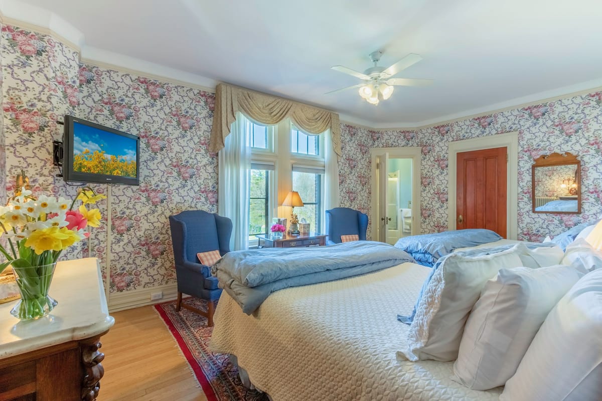 Ein Schlafzimmer mit floralem Wandpapier, einem Bett mit vielen Kissen, einem Schreibtisch mit Blumen in einer Vase und einem Deckenventilator.