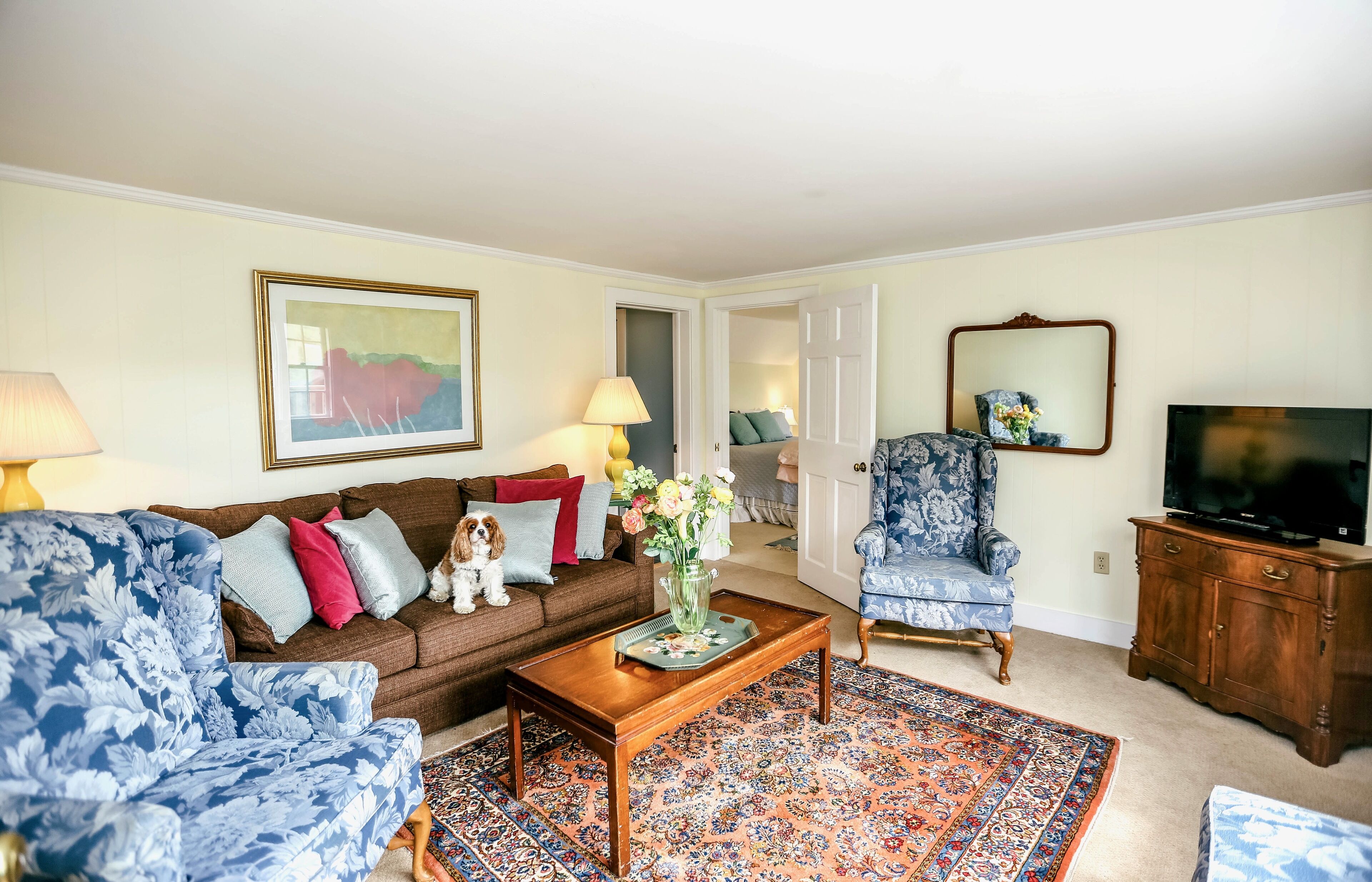 Un salon avec un canapé marron, un fauteuil bleu à motifs floraux, une table basse en bois, un tapis à motifs, un téléviseur et un miroir.