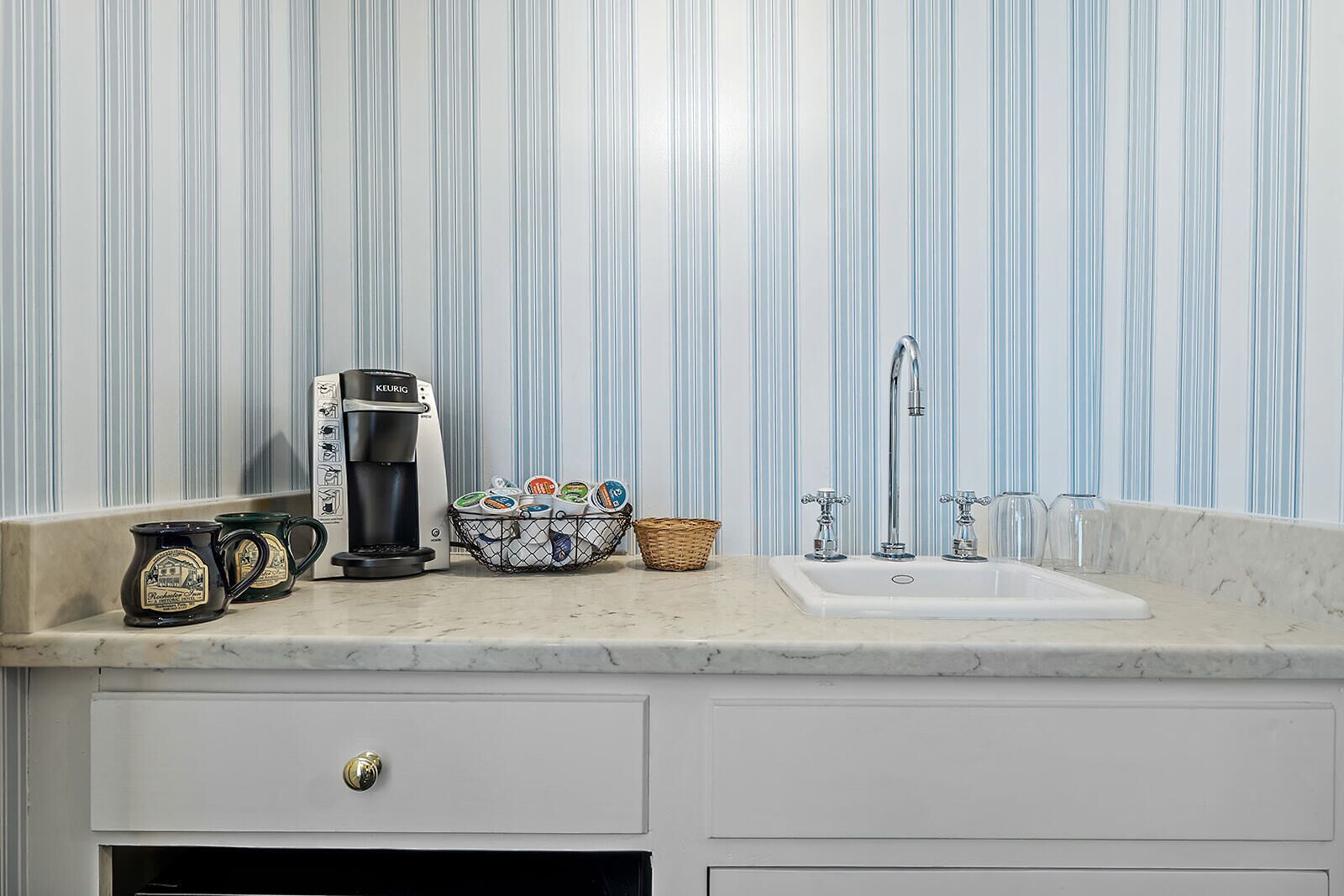 Una cocina con cafetera, tazas, una cesta y un lavabo.