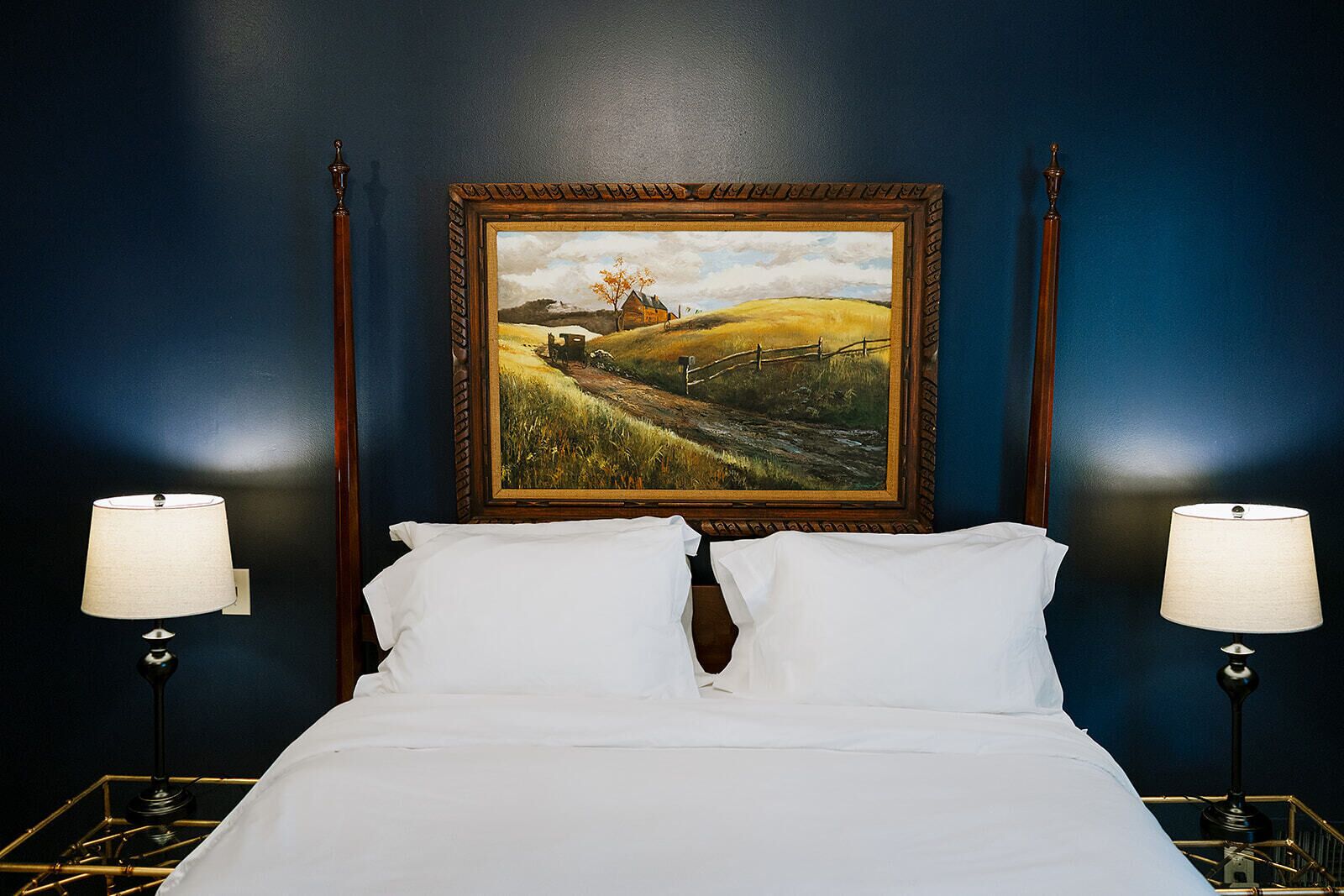 A bed with white linens, two bedside lamps, and a framed landscape painting on the wall.
