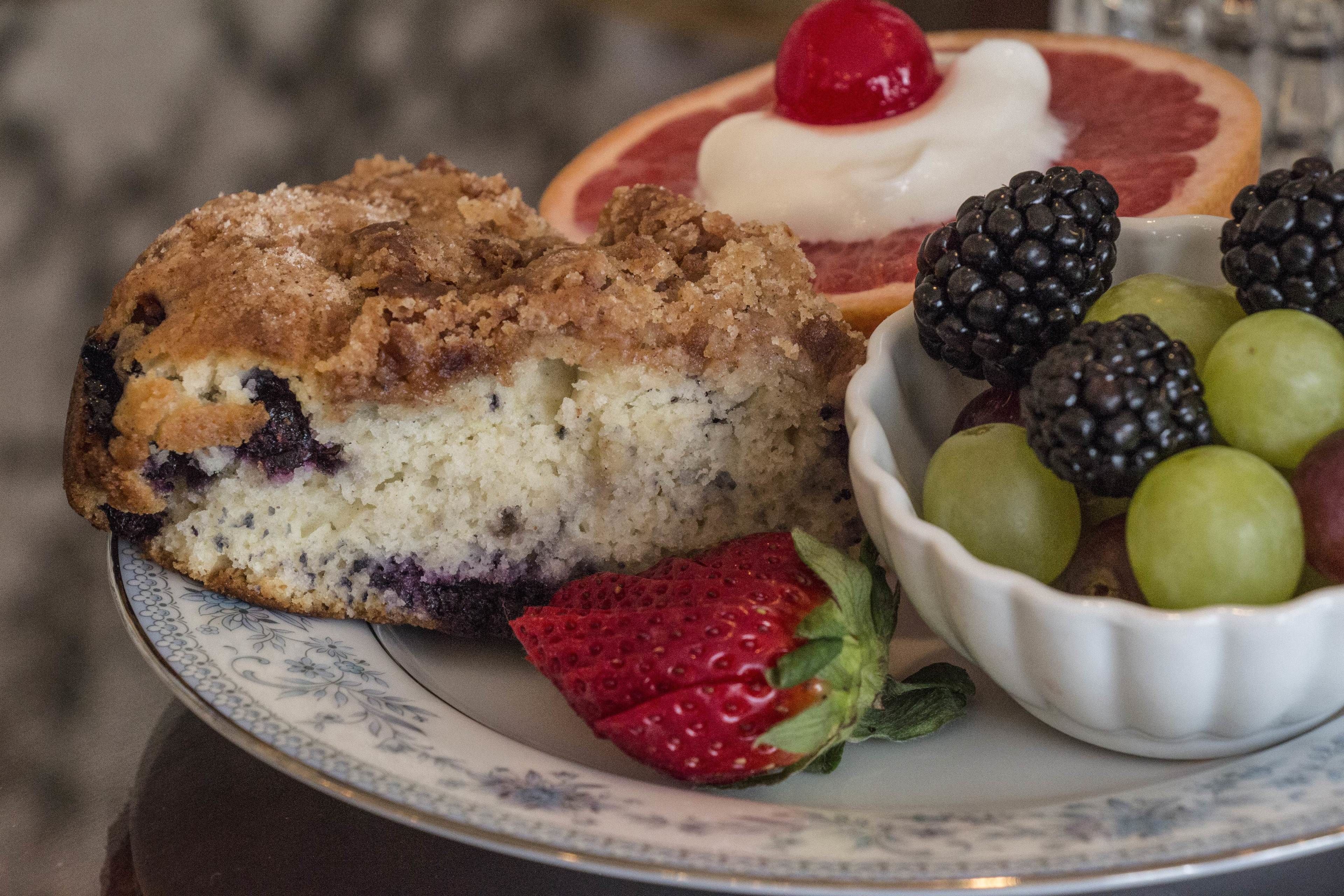 A slice of blueberry muffin with a crumb topping, a slice of grapefruit with whipped cream, and a bowl of mixed berries and grapes.