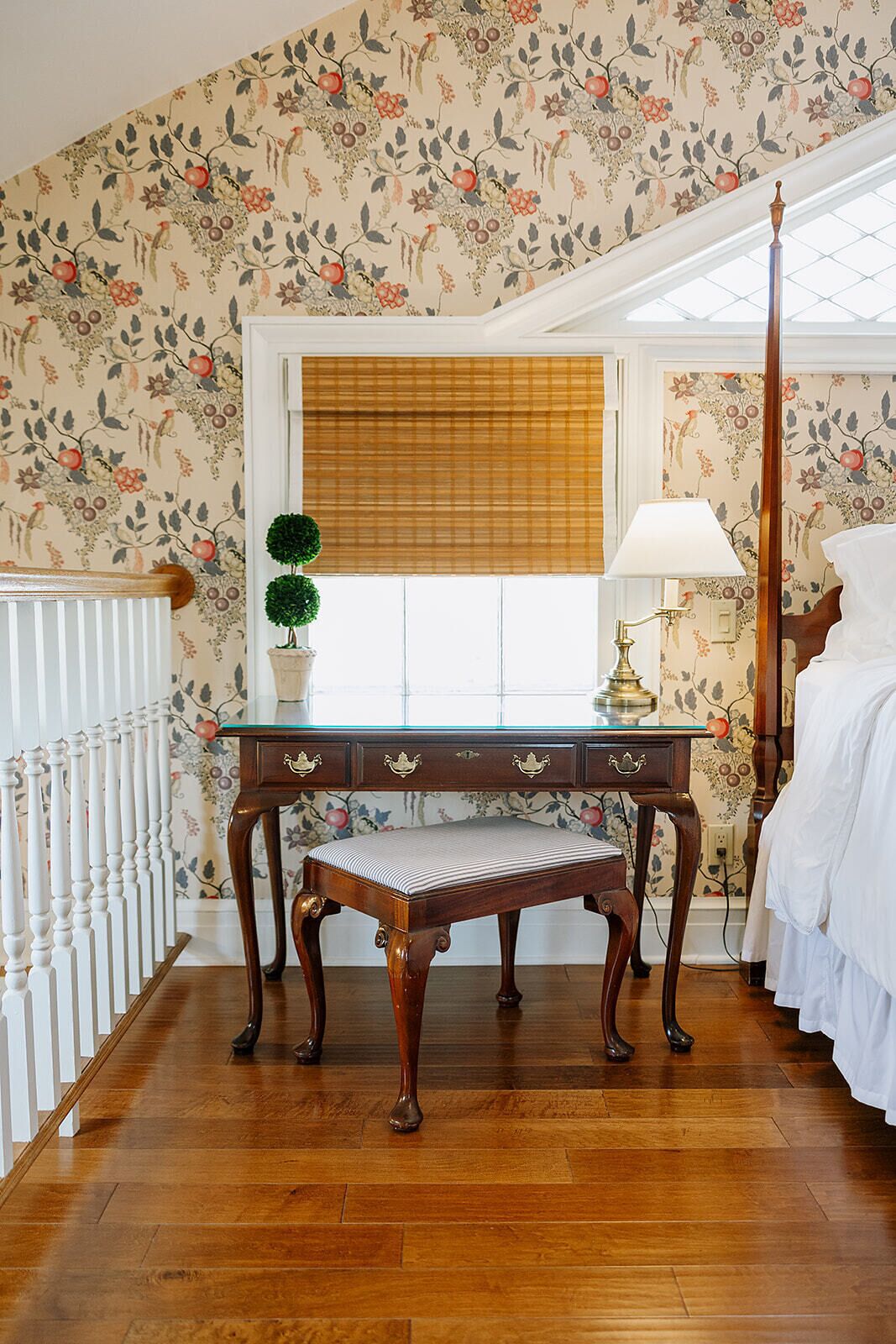 A room with a wooden desk, a bench, a lamp, a window with blinds, and a bed.