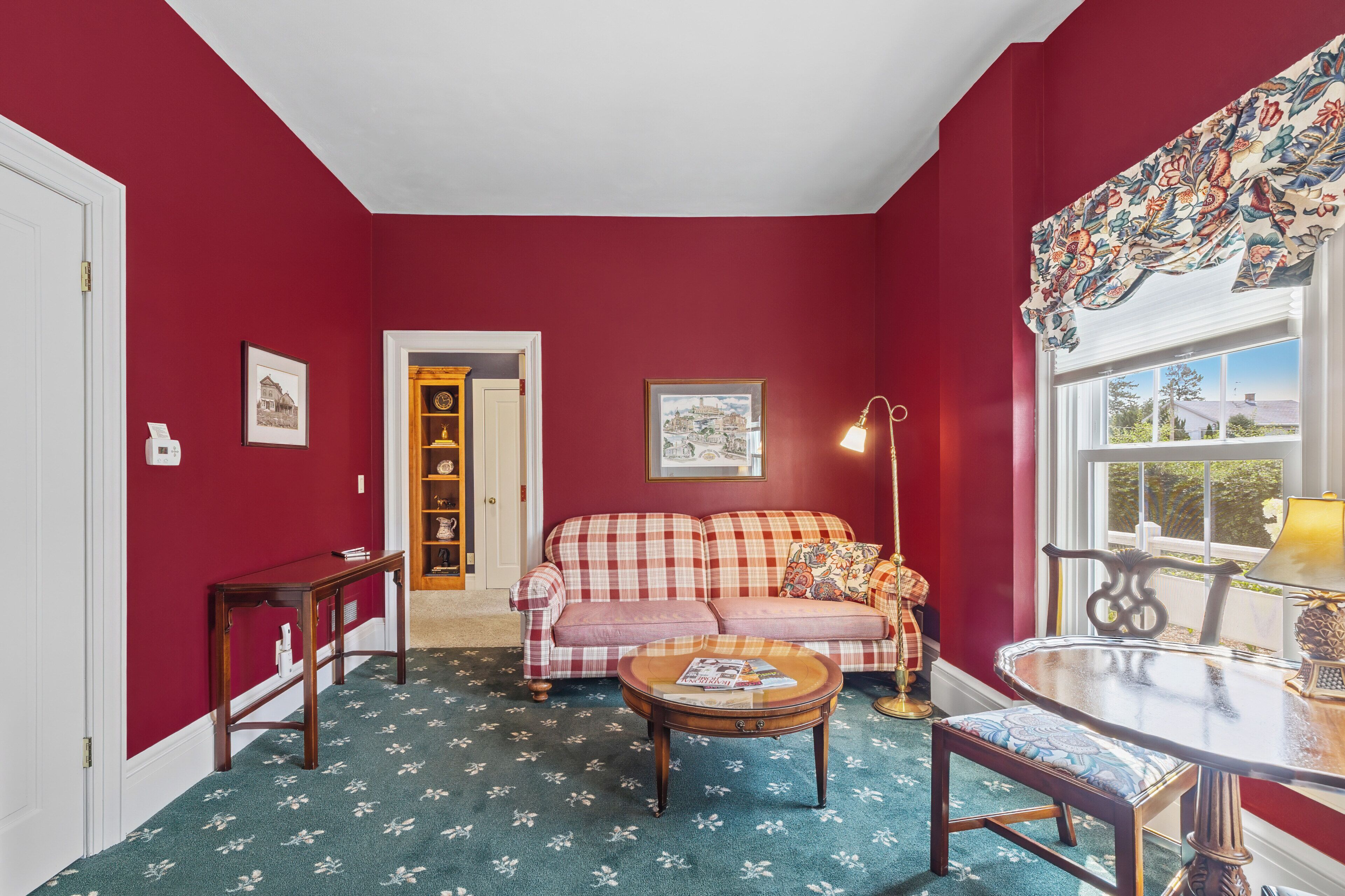 A room with a red wall, a checkered sofa, a round coffee table, a small table with a lamp, and a window with a view.