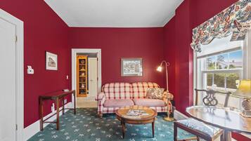 A room with a red wall, a checkered sofa, a round coffee table, a small table with a lamp, and a window with a view.