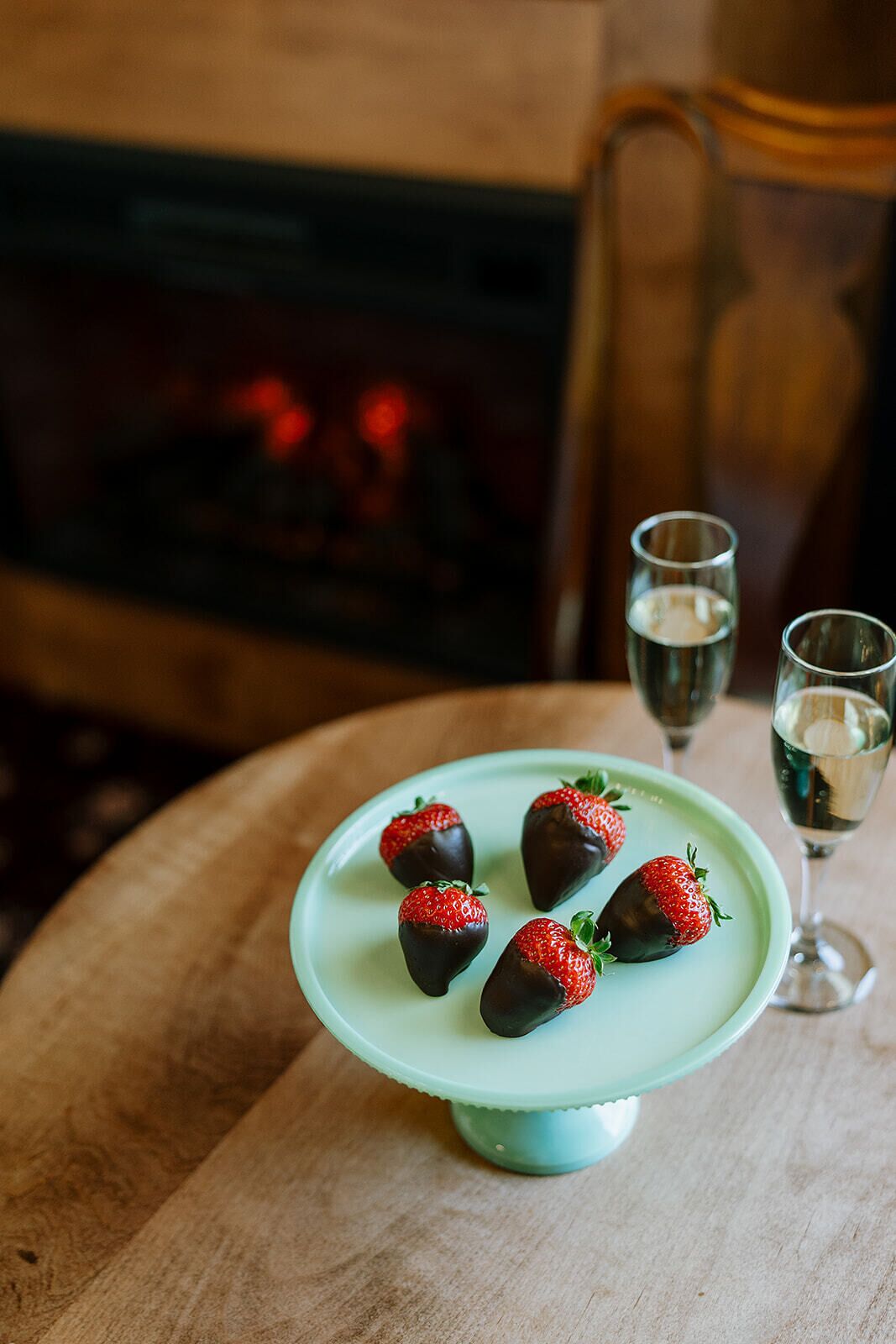 Un plato de fresas cubiertas de chocolate sobre un plato azul claro, una copa de vino y un vaso de agua sobre una mesa de madera con una chimenea al fondo.