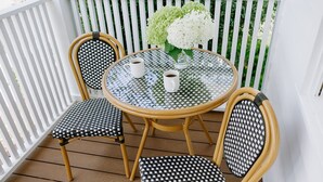 Een ronde glazen tafel met twee stoelen, elk met een zwart-wit geweven zitvlak en rugleuning, en een witte vaas met witte bloemen op de tafel.