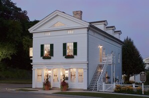 A two-story white building with green shutters, a front porch, and a staircase leading to the upper floor.