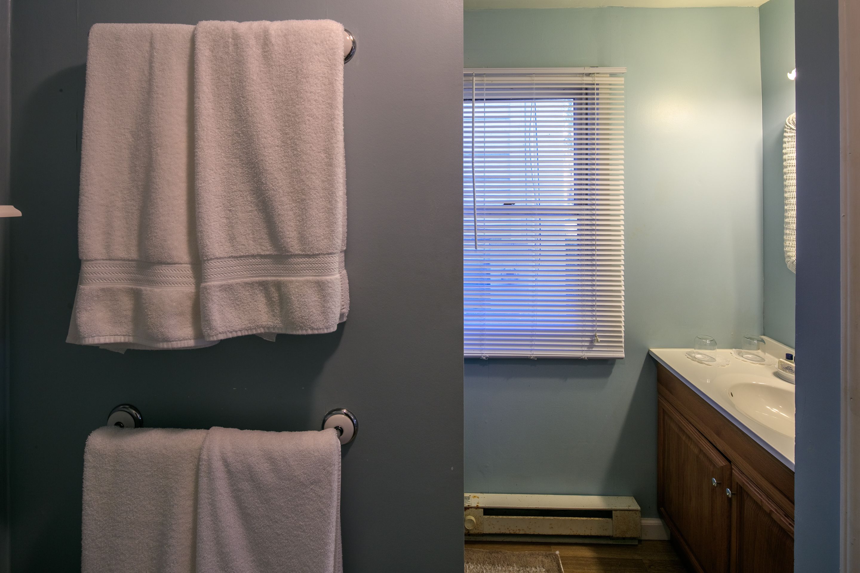 A bathroom with two towels hanging on towel rings, a window with blinds, a sink with a mirror, and wooden cabinets.
