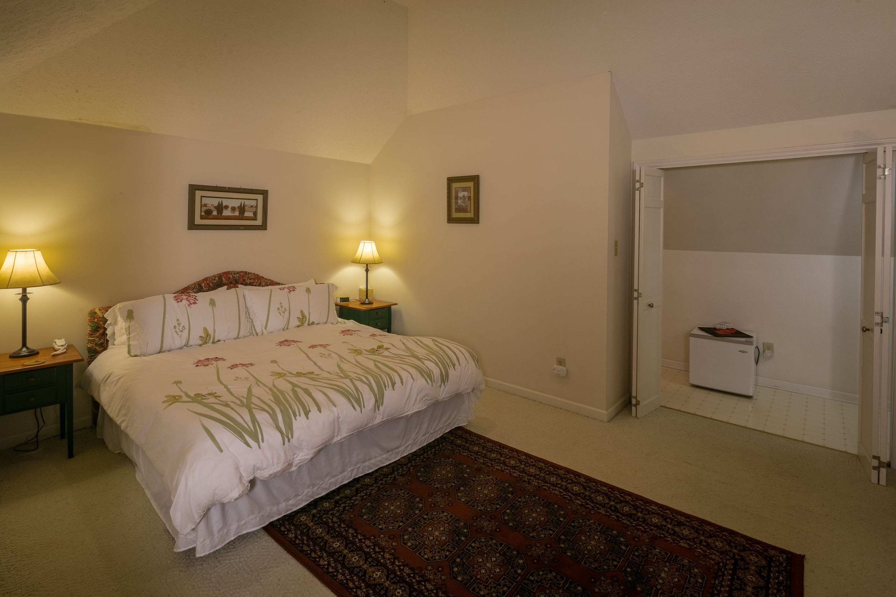 A bedroom with a bed, two bedside lamps, a small bedside table, a rug, and a door leading to another room.
