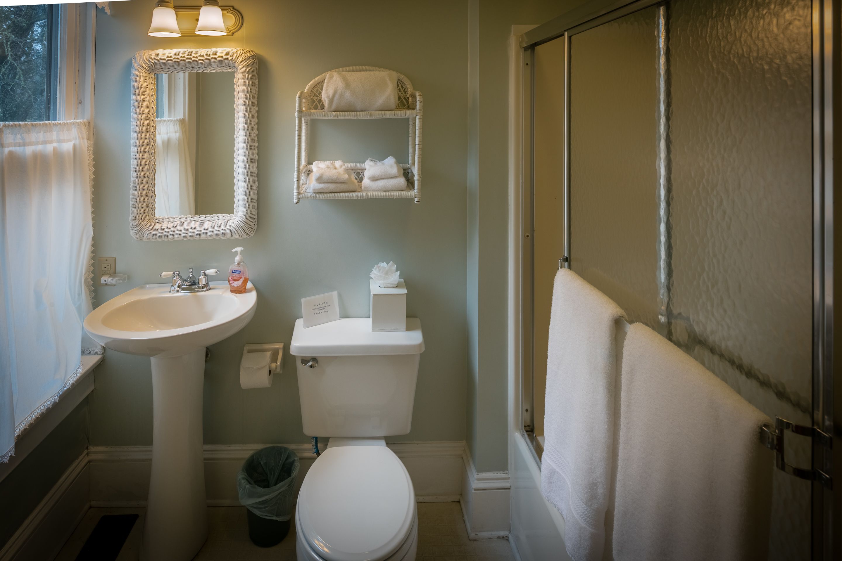 Une salle de bain avec un lavabo sur colonne, des toilettes, une étagère en osier avec des serviettes et une porte de douche.