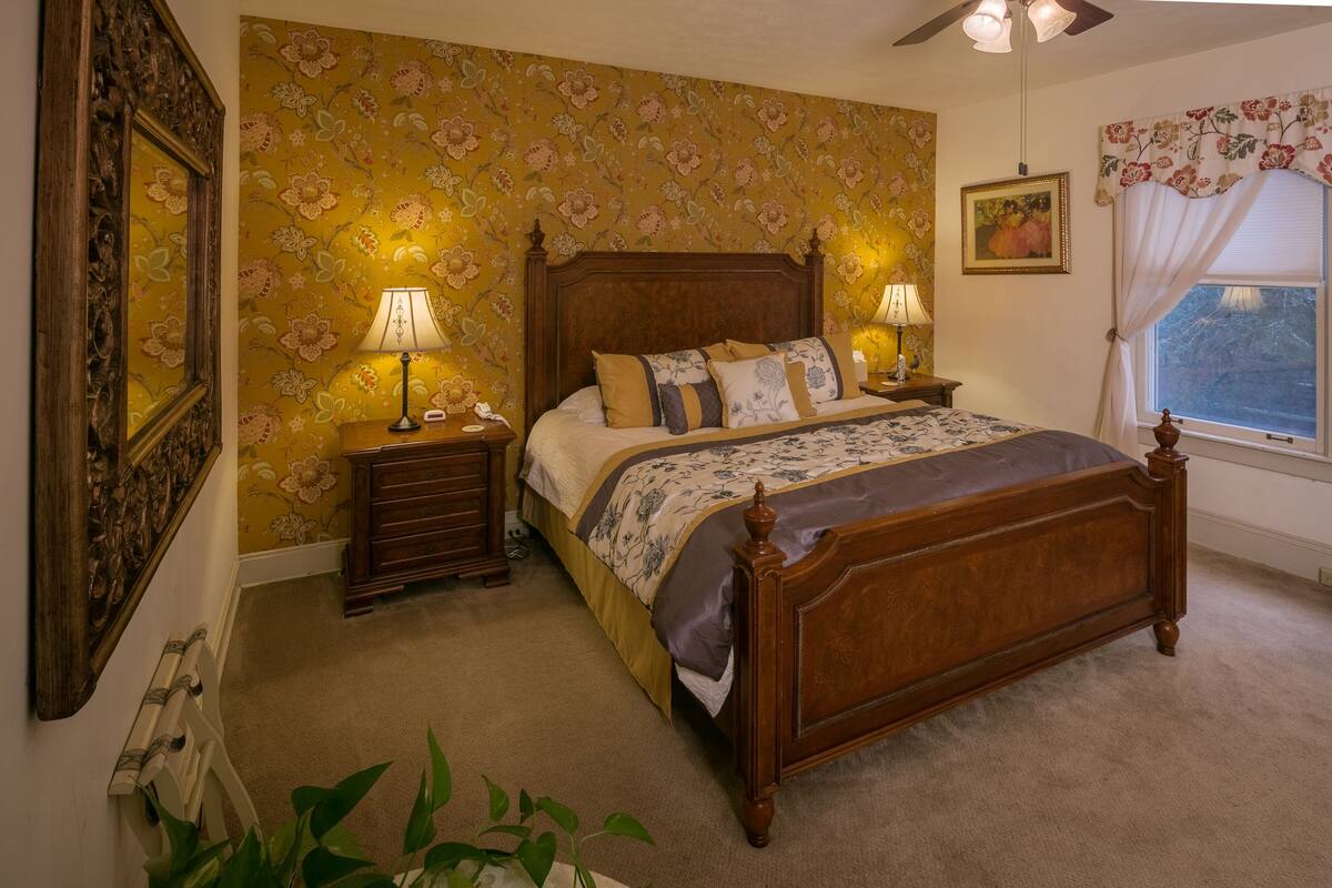 A bedroom with a wooden bed, floral wallpaper, two bedside tables with lamps, a window with curtains, and a framed picture on the wall.
