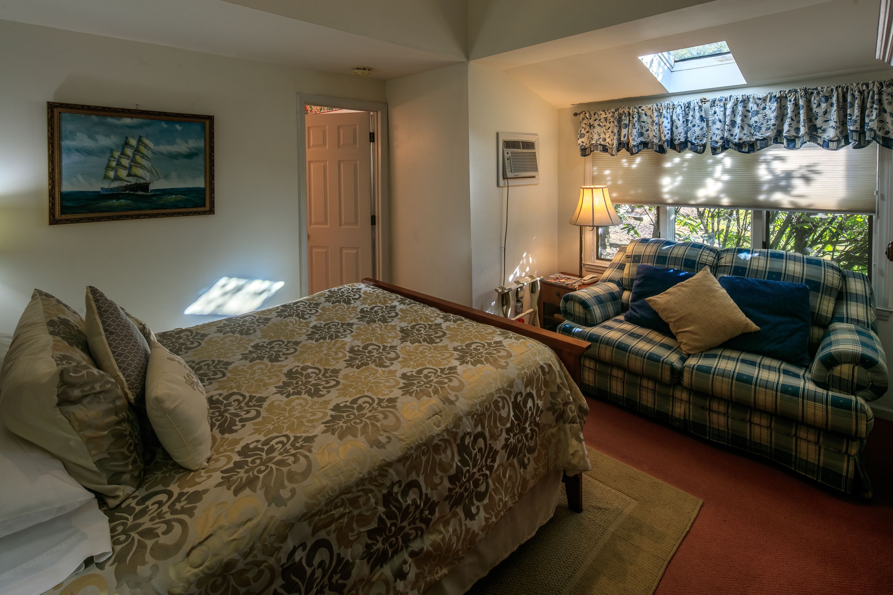 A bedroom with a bed, a sofa, a painting, a window with curtains, and a skylight.