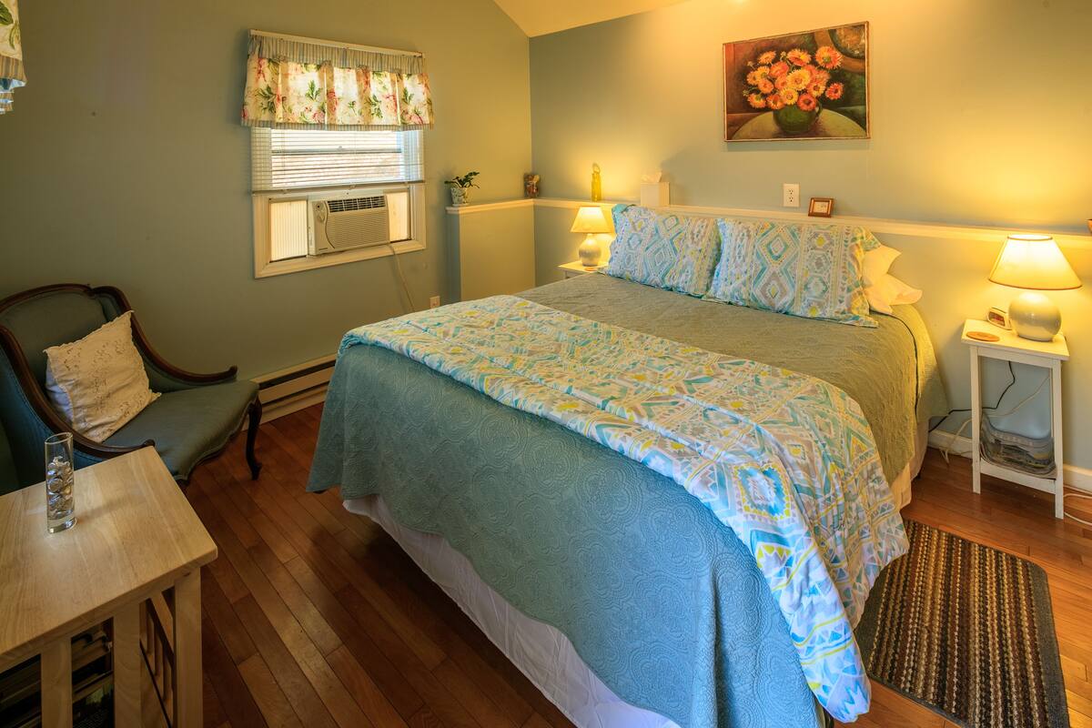 A bedroom with a bed, bedside tables, a chair, a window with blinds, and a painting on the wall.