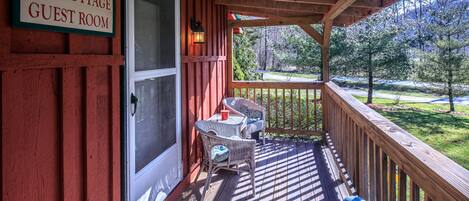 Un porche de madera con un letrero que dice "Red Cottage Guest Room", una puerta, una pequeña mesa con sillas y vistas a un paisaje verde.