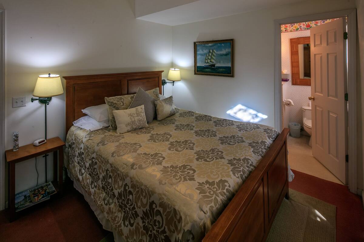 A bedroom with a large bed, two bedside lamps, a nightstand, a magazine, a painting on the wall, and a door leading to a bathroom.