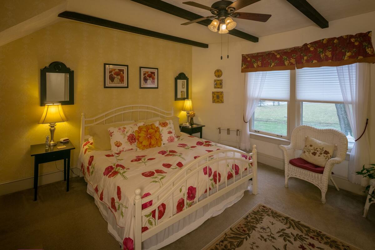 A bedroom with a bed, two bedside tables with lamps, a chair, a window with curtains, and a ceiling fan.