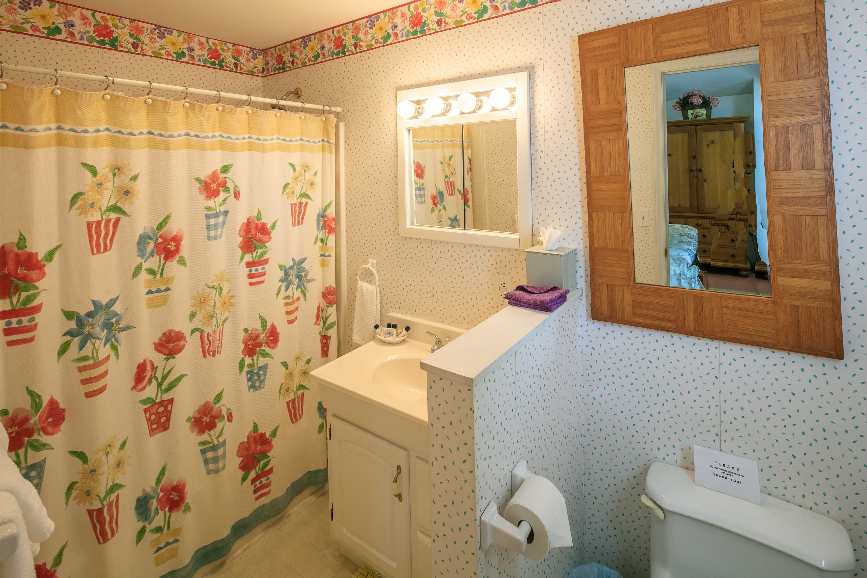 A bathroom with a floral shower curtain, a sink with a mirror above it, and a toilet with a sign on it.