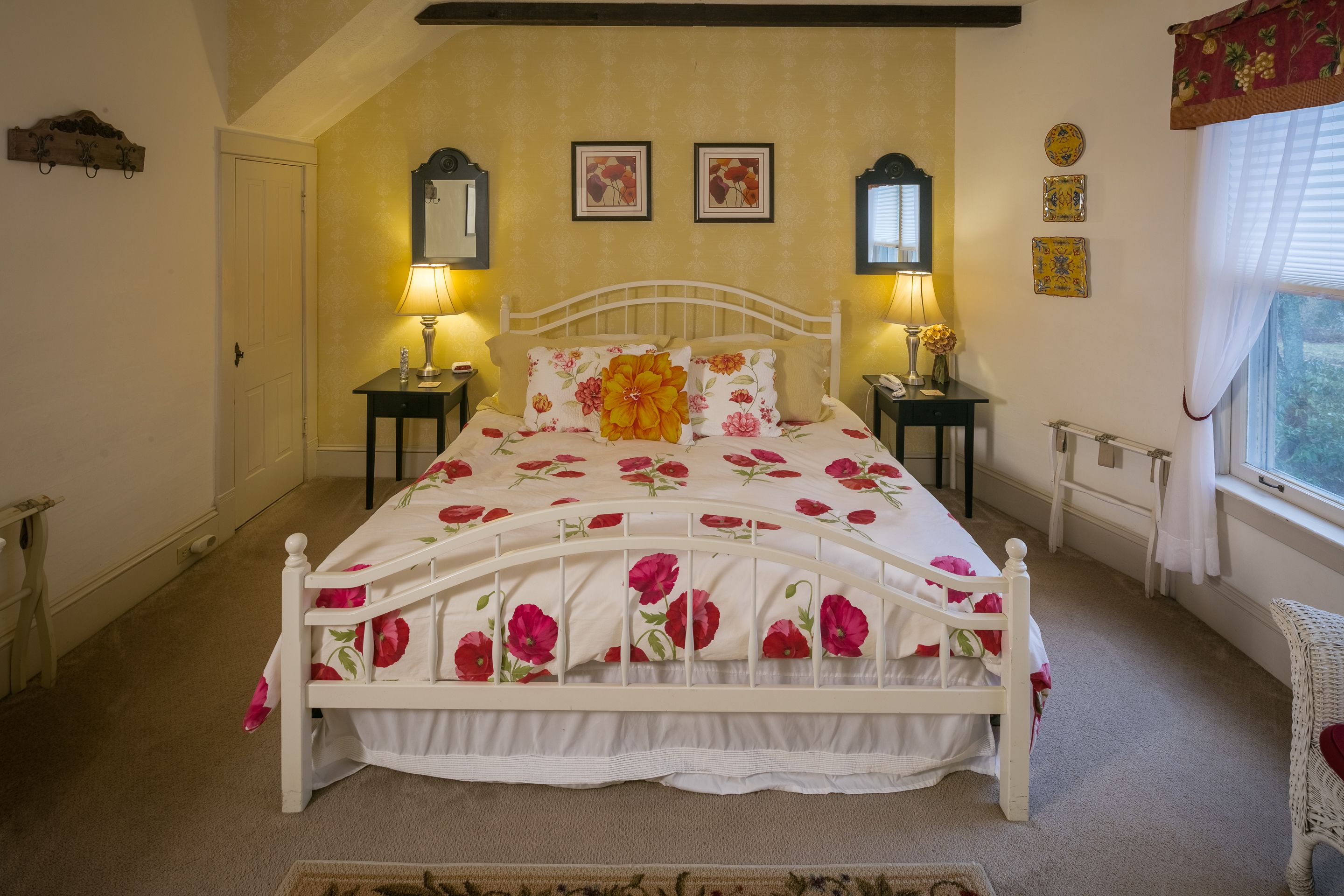 Ein Schlafzimmer mit einem weißen Bett, floralem Bettzeug, zwei Nachttischlampen, einem Fenster mit Vorhängen und einem kleinen Tisch mit einer Lampe.