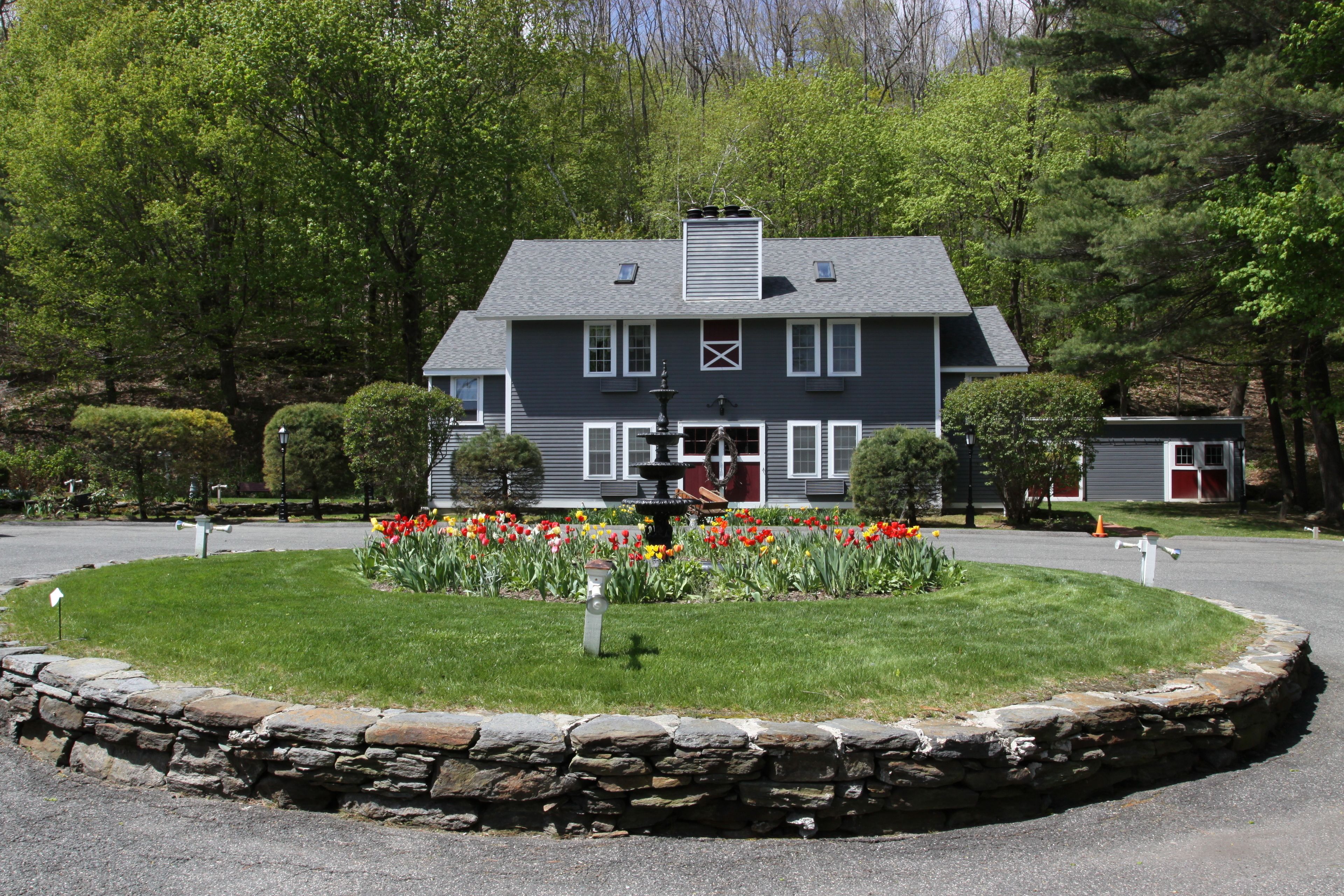 A two-story house with a stone border around the lawn, a fountain, and a variety of flowers.