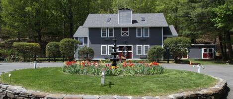 A two-story house with a stone border around the lawn, a fountain, and a variety of flowers.