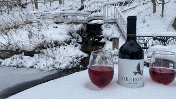 A bottle of Obeton wine and two glasses of red wine on a snow-covered table with a bridge and trees in the background.
