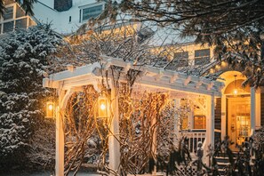 A snow-covered garden with a wooden pergola and lanterns, leading to a building entrance.
