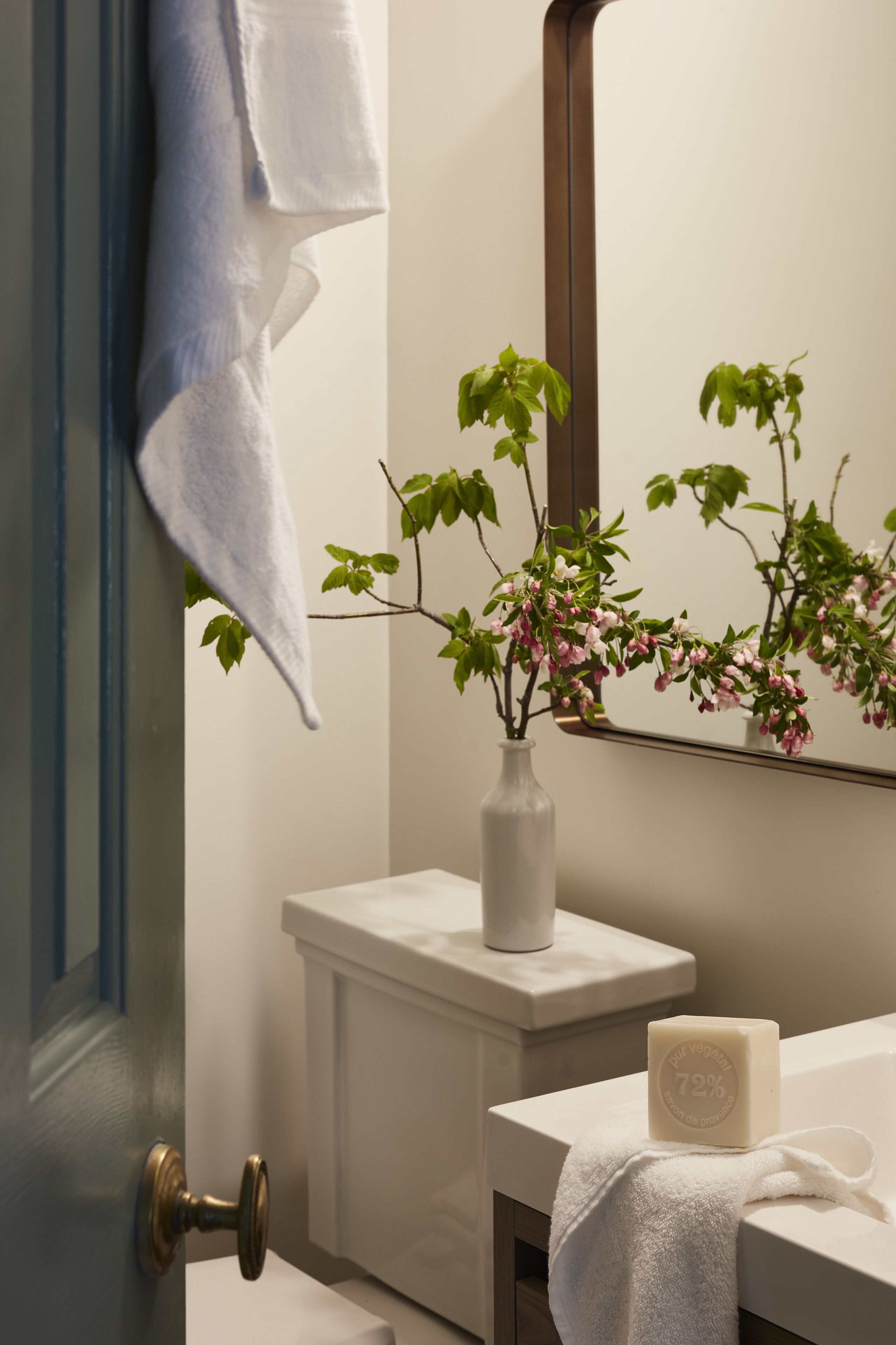 Une salle de bain avec un lavabo blanc, un miroir, un vase de fleurs et une serviette accrochée à la porte.