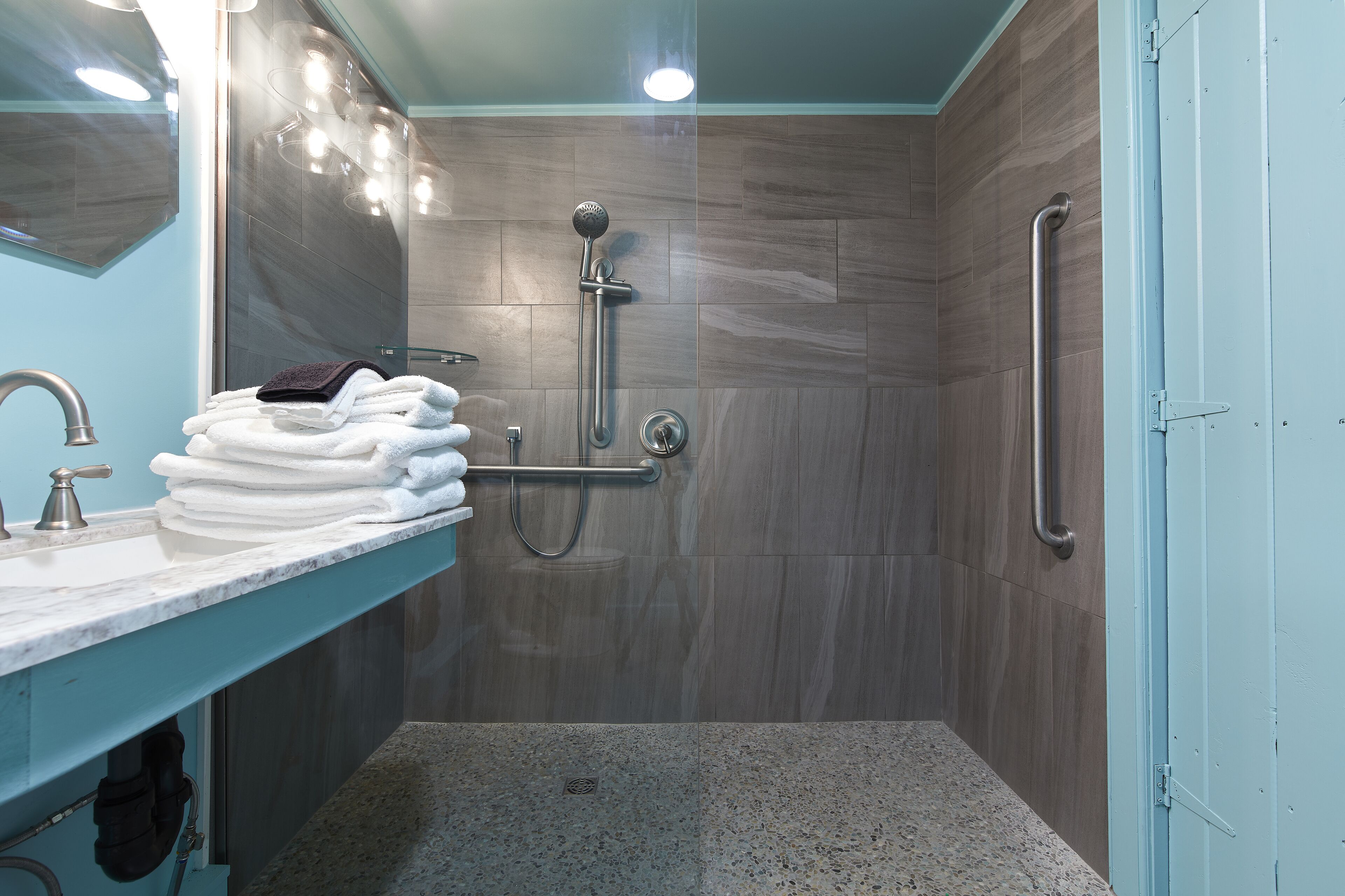 A modern bathroom with a shower, grab bars, a sink with stacked towels, and a mirror.